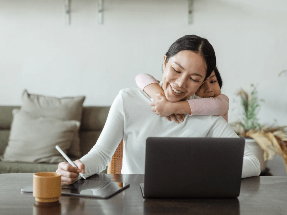 Smiling mother working remotely on a laptop while her young daughter hugs her from behind.