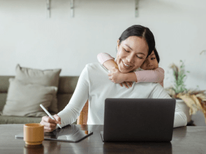 Smiling mother working remotely on a laptop while her young daughter hugs her from behind.